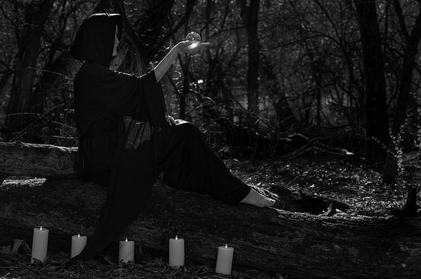 一位身穿黑色长袍、被燃烧的蜡烛包围的妇女正坐在森林里的树干上，手里拿着一个透明的水晶球。巫术。