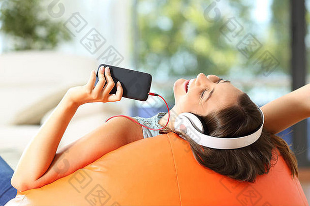 青少年在家里的客厅里，戴着耳机和智能手机躺在橙色的沙发上听音乐