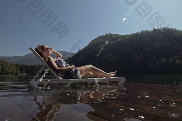 那个女人躺在日光浴床上，戴着太阳镜，披着波西米亚丝绸披肩。女孩在洪水淹没的木头水下码头上休息。湖里的路面被水覆盖着。