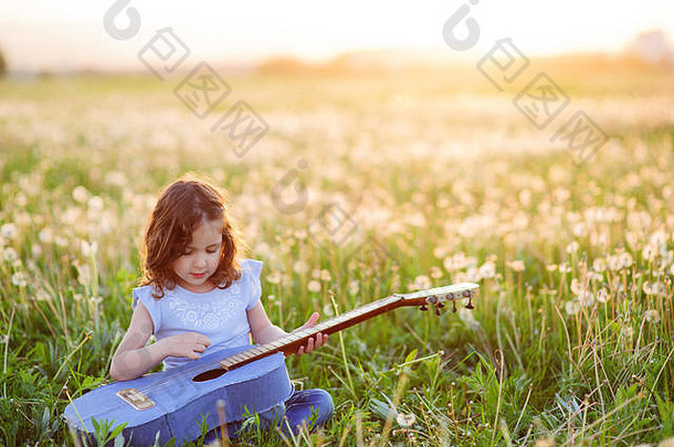 可爱的小卷发女孩坐在夏日田野的绿草上弹吉他
