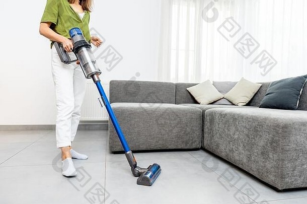 在现代白色客厅里，一位女士用无绳吸尘器清洁地板。使用无线吸尘器轻松清洁的概念