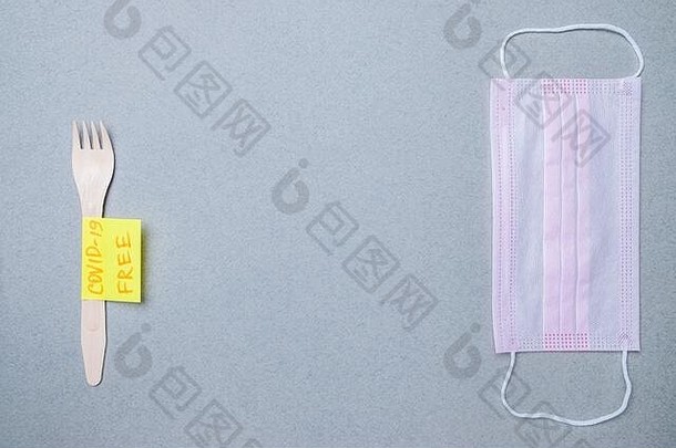 木制叉子，粉红色的医用面具和贴纸，铭文COVID-19免费在灰色背景。提供安全、未受感染的食品的理念