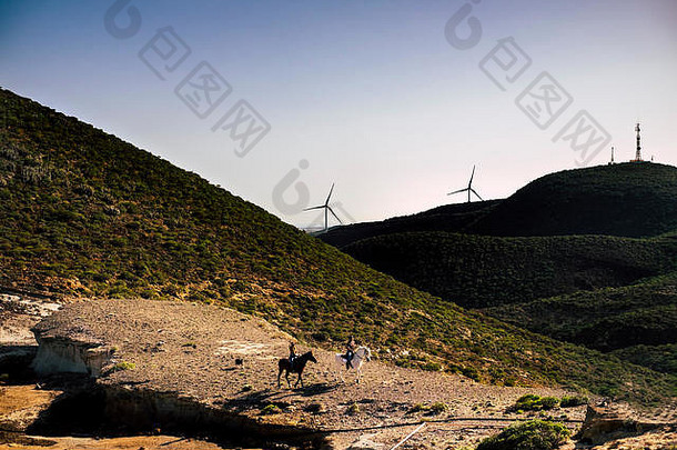 一对骑手带着马在山谷中进行冒险休闲旅游活动，背景是山脉和风力发电厂。自由的