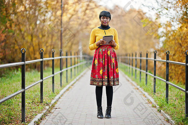 在金秋秋季公园，穿着黄红色连衣裙的非裔美国女孩手持平板电脑。