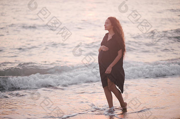 美丽的年轻的怀孕了女人长卷曲的健康的头发穿黑色的衣服走海滩日落未来妈妈。概念