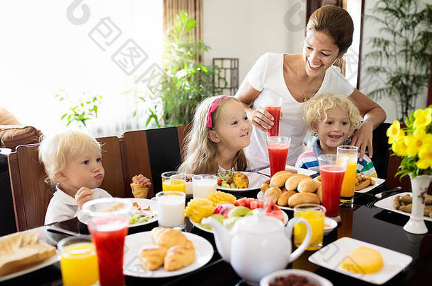 在家吃健康的家庭早餐。母亲和孩子们吃热带水果、烤面包、奶酪和香肠。孩子们在阳光充足的天气喝新鲜的榨果汁