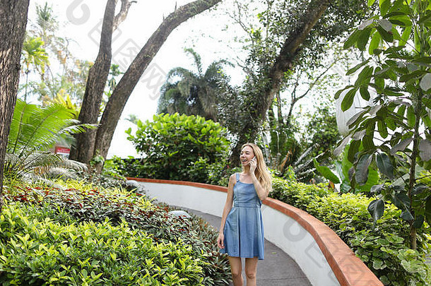 穿着牛仔裤和太阳裙的年轻女子在热带植物附近通过智能手机聊天。