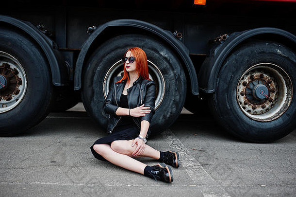红头发的时髦女孩穿着黑色衣服，靠着大卡车车轮坐着。