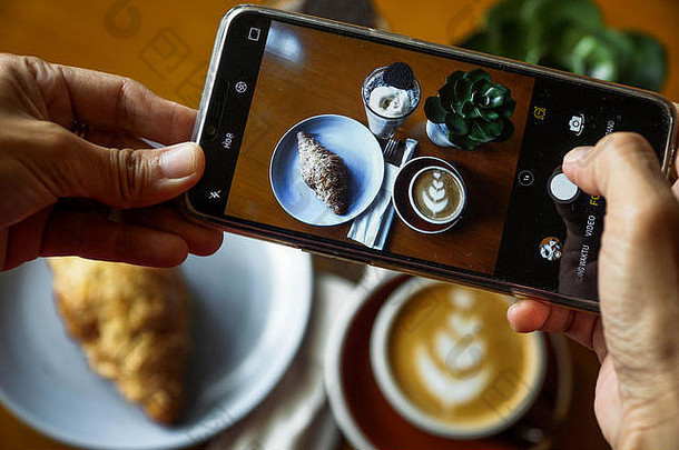 用手机拍摄食物。咖啡拿铁、牛角面包巧克力和蓝色海洋的产品照片。一家咖啡馆的食物俯视图
