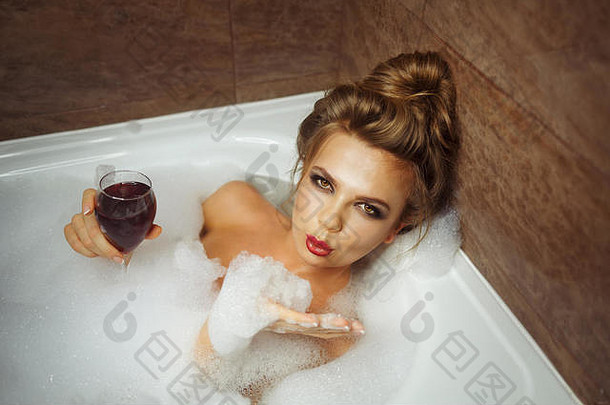 年轻可爱的女孩在浴池里用泡沫喝红酒。辛苦一天后她放松了下来。女孩风骚地抬起头，送上一个飞吻。