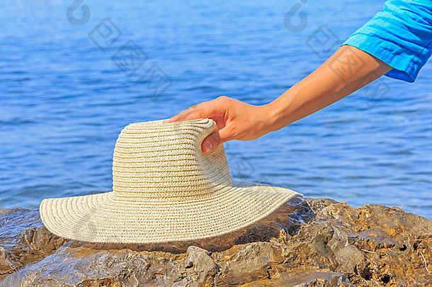 在蓝色海洋衬托下的海滩上，一名女子手持帽子