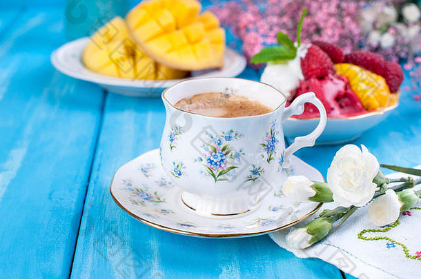 香浓的咖啡装在一个漂亮的瓷杯里，上面有蓝色的花朵和水果味的冰淇淋。蓝色木质背景上的花朵和成熟芒果。早餐很甜