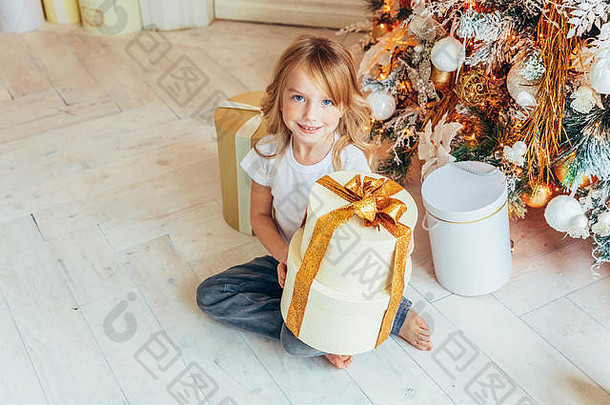 圣诞前夜，小女孩在家里的圣诞树旁拿着礼品盒。孩子们在有冬季装饰的明亮房间里。家庭幸福。圣诞节-新年-十二月-庆祝时间概念。