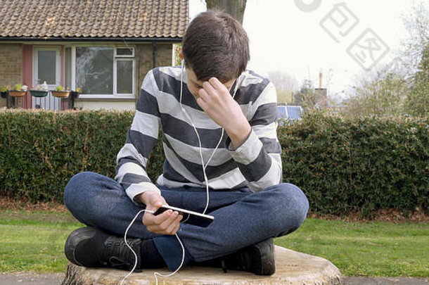 坐在英国公园的树桩上用智能手机听或说的十几岁男孩。青少年与社交媒体概念。