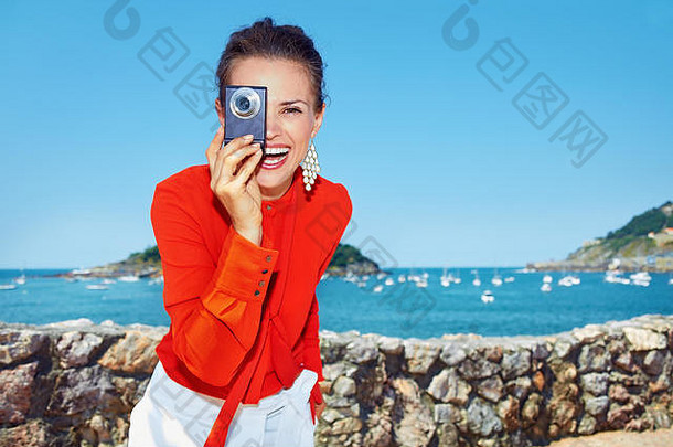 豪华周末度假酒店。穿着鲜艳上衣的快乐年轻女子，站在美丽的风景前，用游艇俯瞰环礁湖，假装用数码相机拍照