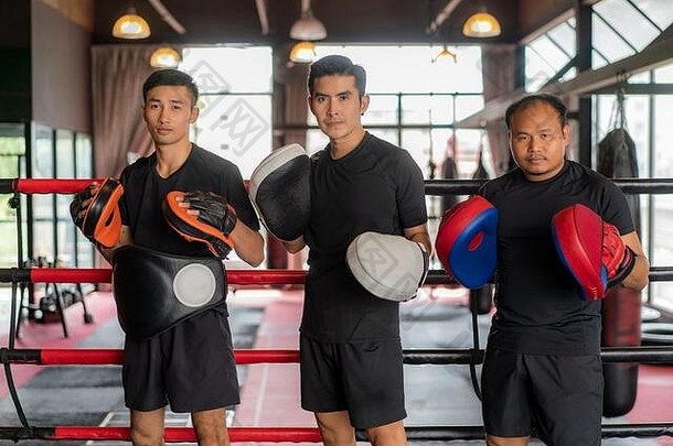 三名亚洲拳击手在拳击台上靠着黑红色的绳子，看着摄像机摆出姿势，微笑着，在blac艰苦训练后休息
