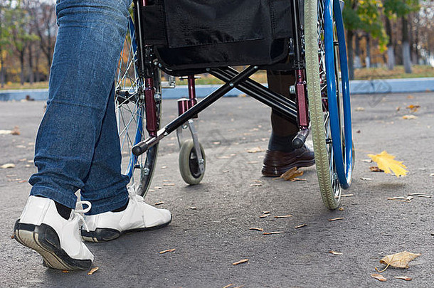 一个穿着牛仔裤和运动鞋的看护者推着轮椅沿着柏油路的双腿特写镜头，低角度视角