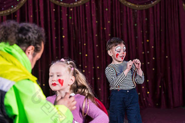 男孩扮成小丑，化妆，穿着条纹衬衫，在舞台上用红窗帘表演，女孩在脸上化妆