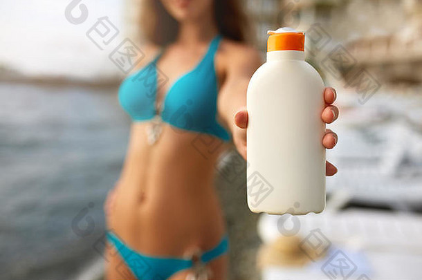 快乐女人显示晒黑防晒霜奶油白色瓶模糊海滩背景晒黑苗条的女孩穿蓝色的比基尼泳衣持有