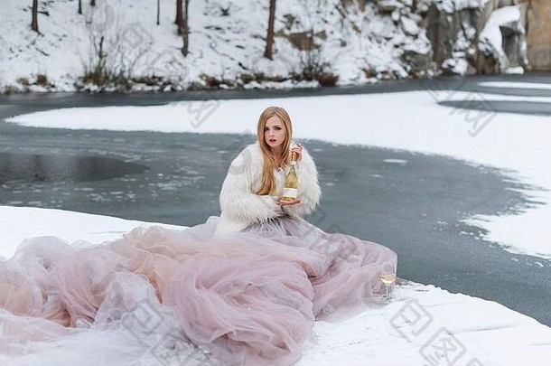 冬天在户外喝酒的金发女人。穿着长袍的时装模特坐在冰雪覆盖的湖面上喝酒庆祝。冬天