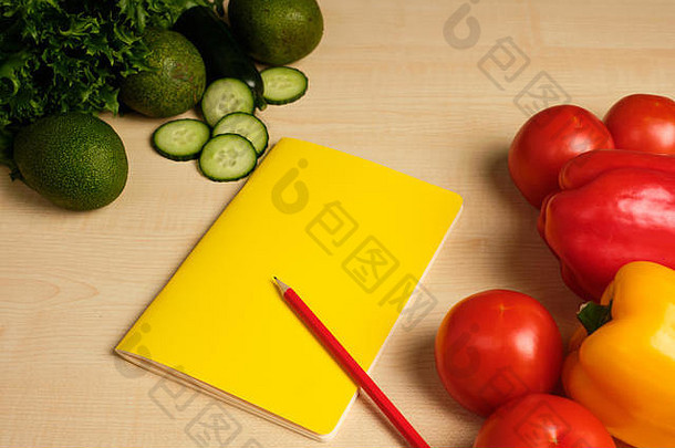 绿色和红色的蔬菜在木桌上相对放置，中间有一个黄色的记事本。