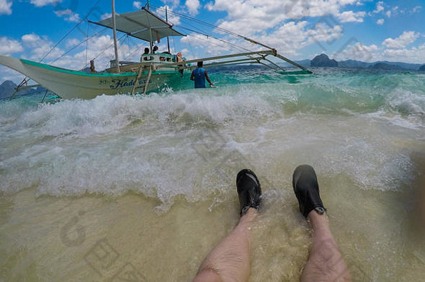 巴拉望岛El Nido——2019年2月26日——男子双腿在海滩浪花中的旅行照片，背景是小船。