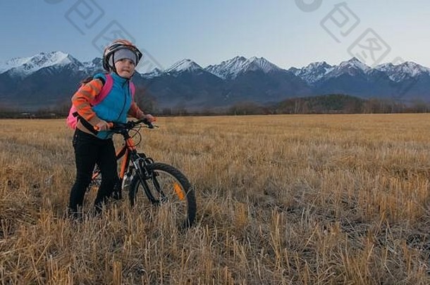 一个白人孩子在麦田里骑自行车。小女孩在美丽的雪山背景下走着黑橙色的自行车。自行车看台