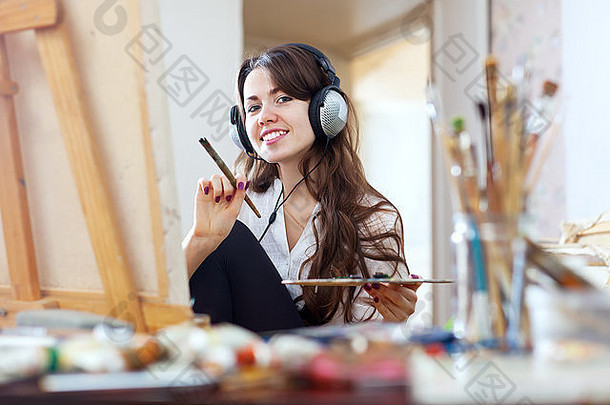 戴着耳机的长发女孩用油画颜料和画笔在画布上作画
