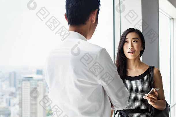 一位商人和女商人站在一扇大窗户旁交谈，可以俯瞰一座城市