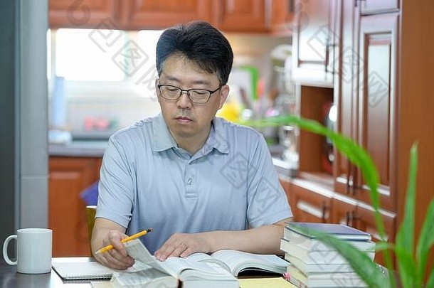 一位亚洲中年男子坐在家里的书桌旁，读书和学习。