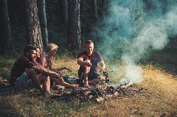 两对夫妇在树林里露营。在篝火上煮香肠。森林里温暖的夏夜