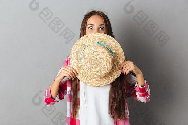 一位年轻有趣的女士站在灰色背景下，将脸藏在一顶帽子后面的肖像