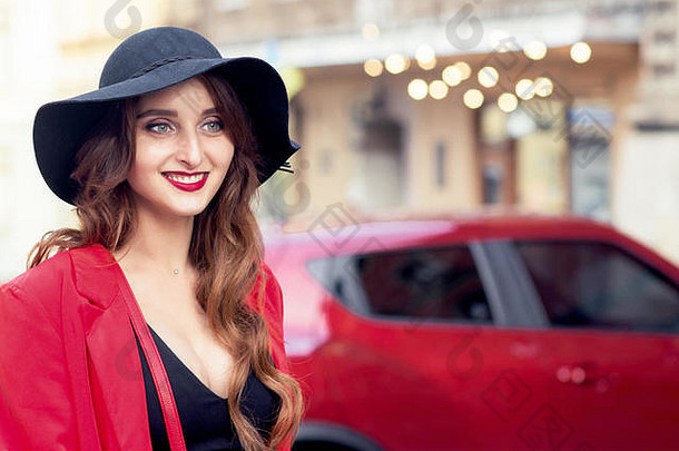 红色汽车旁戴着黑帽子、面带微笑的客户妇女。红色汽车背景上戴黑帽子的年轻女子。