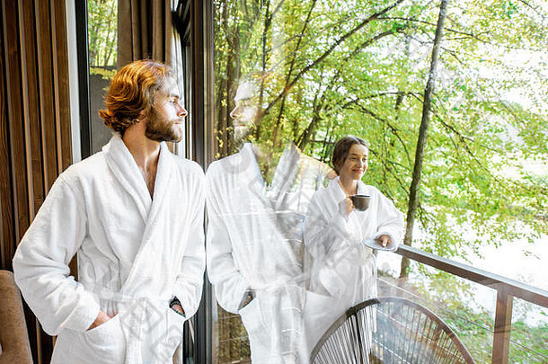 这对穿着浴衣的年轻夫妇站在现代酒店房间的窗户旁，可以看到森林的美丽景色