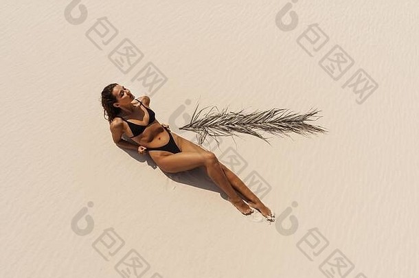上图是一位身穿泳衣的妇女在海边放松和日光浴的空中无人机照片。穿着黑色比基尼的迷人黑发女孩躺在沙滩上，身上长着棕榈叶。美丽的棕褐色混血亚裔白人度<strong>假模特</strong>。暑期时尚概念