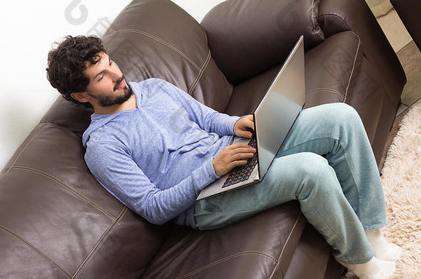 成人在客厅里上网搜索房价。在家里，在客厅里。穿着蓝色和浅绿色睡衣，留着胡子的男人。高昂