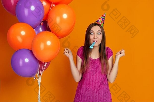 穿着粉色t恤、戴着生日帽、吹着派对号角的鲜艳彩色气球的快乐有趣的年轻女子。