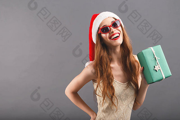 戴着红色圣诞老人帽子和红唇太阳镜的快乐兴奋的女人微笑着，拿着绿色包装的礼物，她站在灰色背景上，新年、圣诞节、假期、纪念品、礼物、购物、折扣、商店、雪人圣诞老人、化妆、发型、狂欢节。