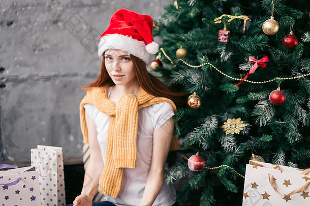 一位留着红色长发的美丽年轻女子在圣诞树旁休息，看着相机，特写照片。美貌、<strong>喜事</strong>