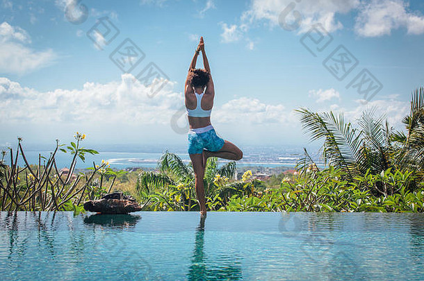 在巴厘岛的别墅里，一位正在练习瑜伽的年轻女子在无限泳池边摆出ashtanga hatha asana的姿势，景色令人叹为观止