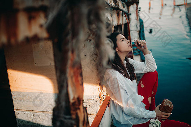 一位年轻女子坐在一艘废弃的旧船上吃面包，她穿着白色衬衫、帽子和红色多孔紧身衣。