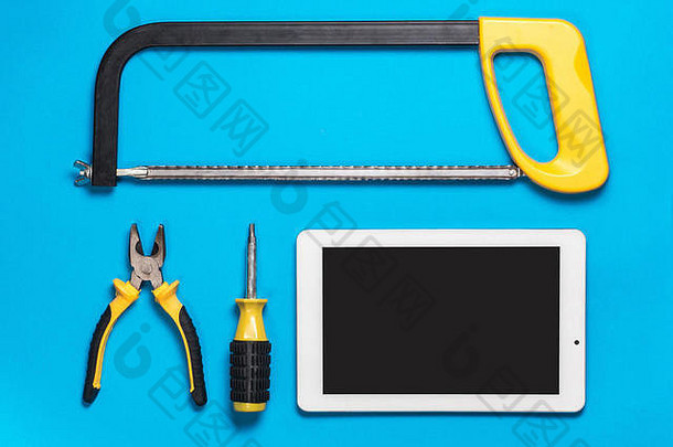 白色的平板和一套黄色的建筑工具在蓝色的桌子上修理：锯子，螺丝刀，钳子。顶视图。机械工作区。