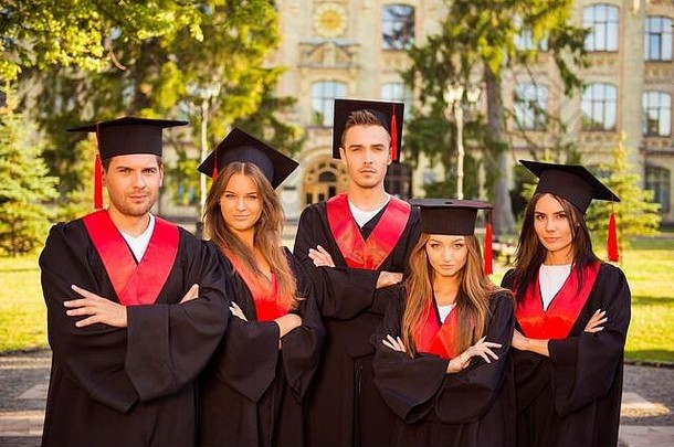 成功自信的五名毕业生穿着长袍，戴着帽子，站成一排，双手交叉