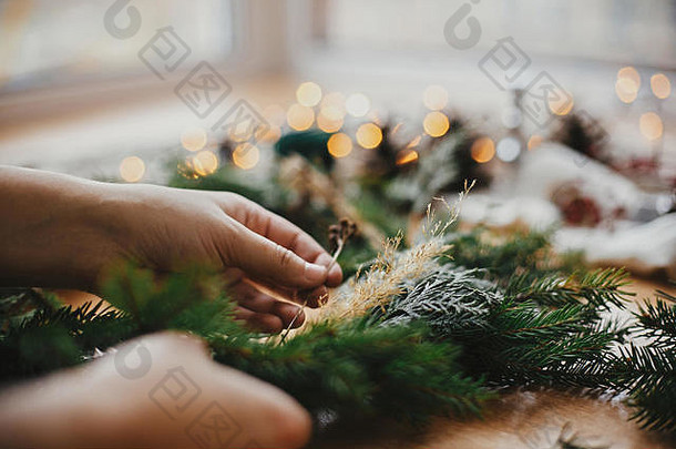 手拿着药草和冷杉枝，松果，线，浆果，木制桌子上的金色灯光。圣诞花圈工作坊。正宗时尚的still l