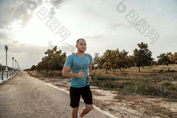 身着蓝色t恤和黑色短裤的白人男子的肖像，日落时在柏油跑道上训练和跑步