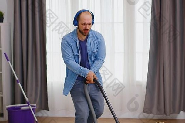 男人一边用耳机听音乐，一边用吸尘器打扫公寓的地板。