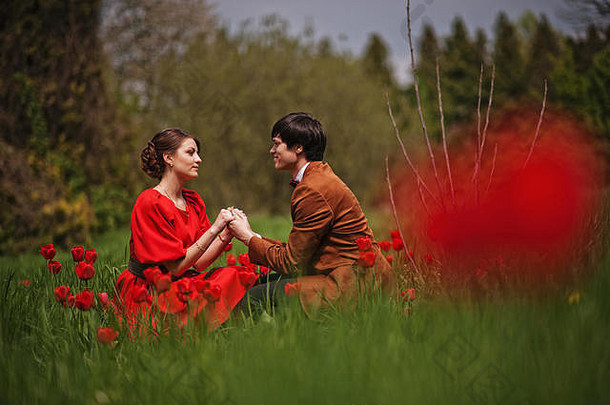 这对情侣在红色郁金香花圃里拥抱爱情。穿着天鹅绒夹克的时髦男人和穿着红色连衣裙的女孩相爱在一起