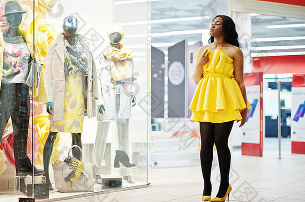 穿着黄色睡衣的时尚非洲裔美国妇女在商场里与模特儿对着陈列柜摆姿势。