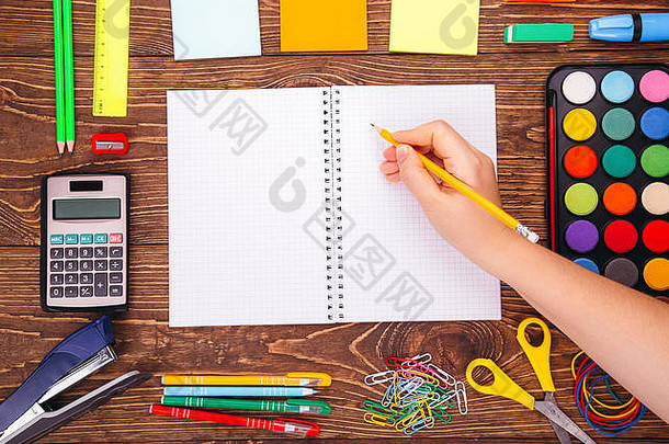 打开空白笔记本，手<strong>拿毛笔</strong>，在木制书桌的背景上放着一个学校用品的框架。将拷贝空间返回到sch