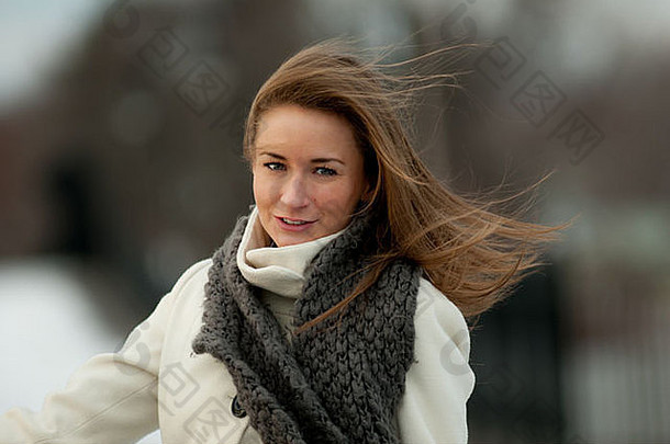 一位穿着白色外套、戴着灰色围巾的美丽年轻女子在刮风的冬日微笑着。